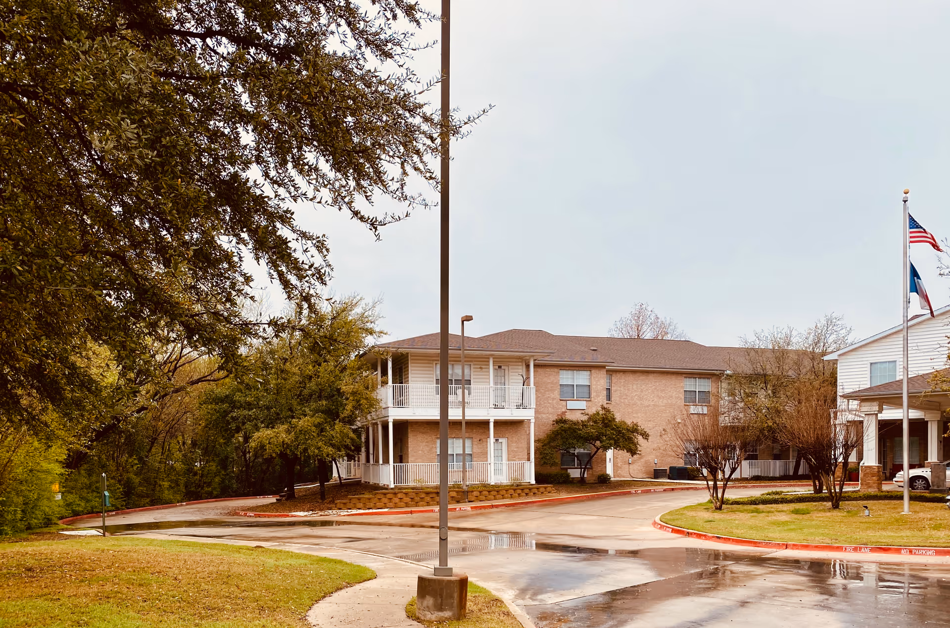 Exterior view of a senior living facility building with a two-story section featuring white railings and balconies. The building is surrounded by trees and a curved driveway with a wet surface. There are two flagpoles with the American and Texas flags near the entrance.