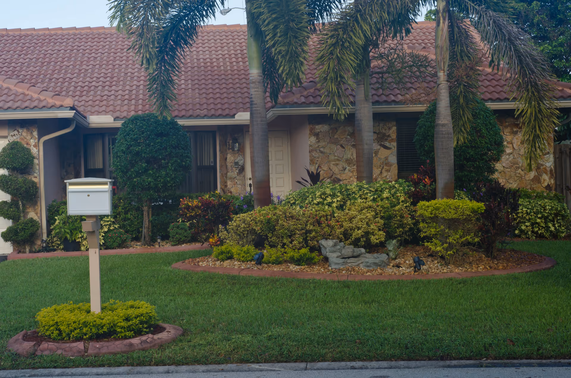 Front view of a single-story house with a tiled roof, stone facade, and a well-maintained garden featuring trimmed bushes, palm trees, and a white mailbox on a post surrounded by greenery.