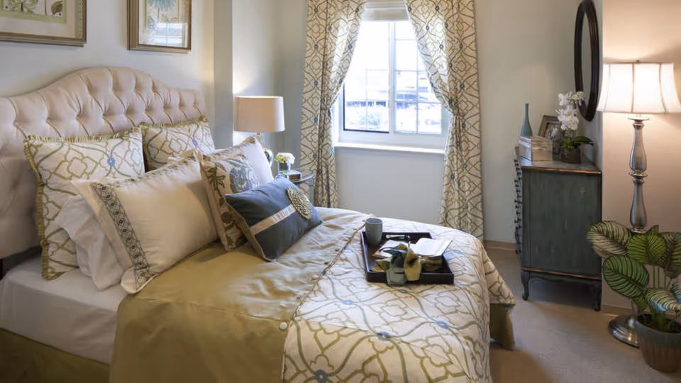 A neatly made bed with multiple decorative pillows and a patterned bedspread in a bright bedroom. There is a window with patterned curtains letting in natural light. A bedside table with a lamp and flowers is on one side, and a small dresser with a round mirror, lamp, and plants is on the other side.