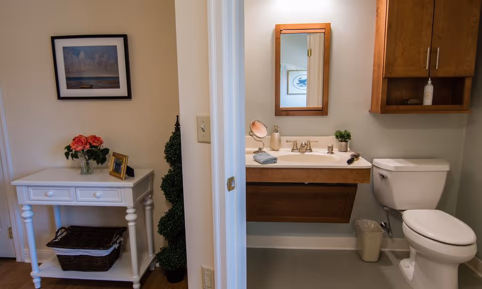 A small bathroom with a white toilet, a wooden vanity with a sink, a mirror above the sink, and a wooden cabinet mounted on the wall. Outside the bathroom, there is a white table with two drawers, a basket underneath, a vase with pink flowers, a framed photo, and a framed picture hanging on the wall above the table.