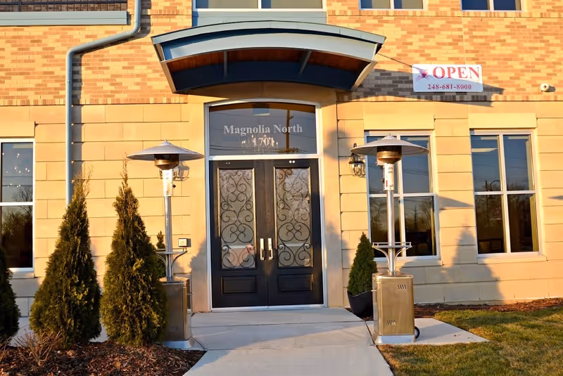 Exterior view of the entrance to a building named Magnolia North with double black doors featuring decorative glass panels, flanked by two outdoor patio heaters and small evergreen shrubs. The building facade is made of light-colored stone and brick, with several windows and a sign above the door that reads 'OPEN' with a phone number.