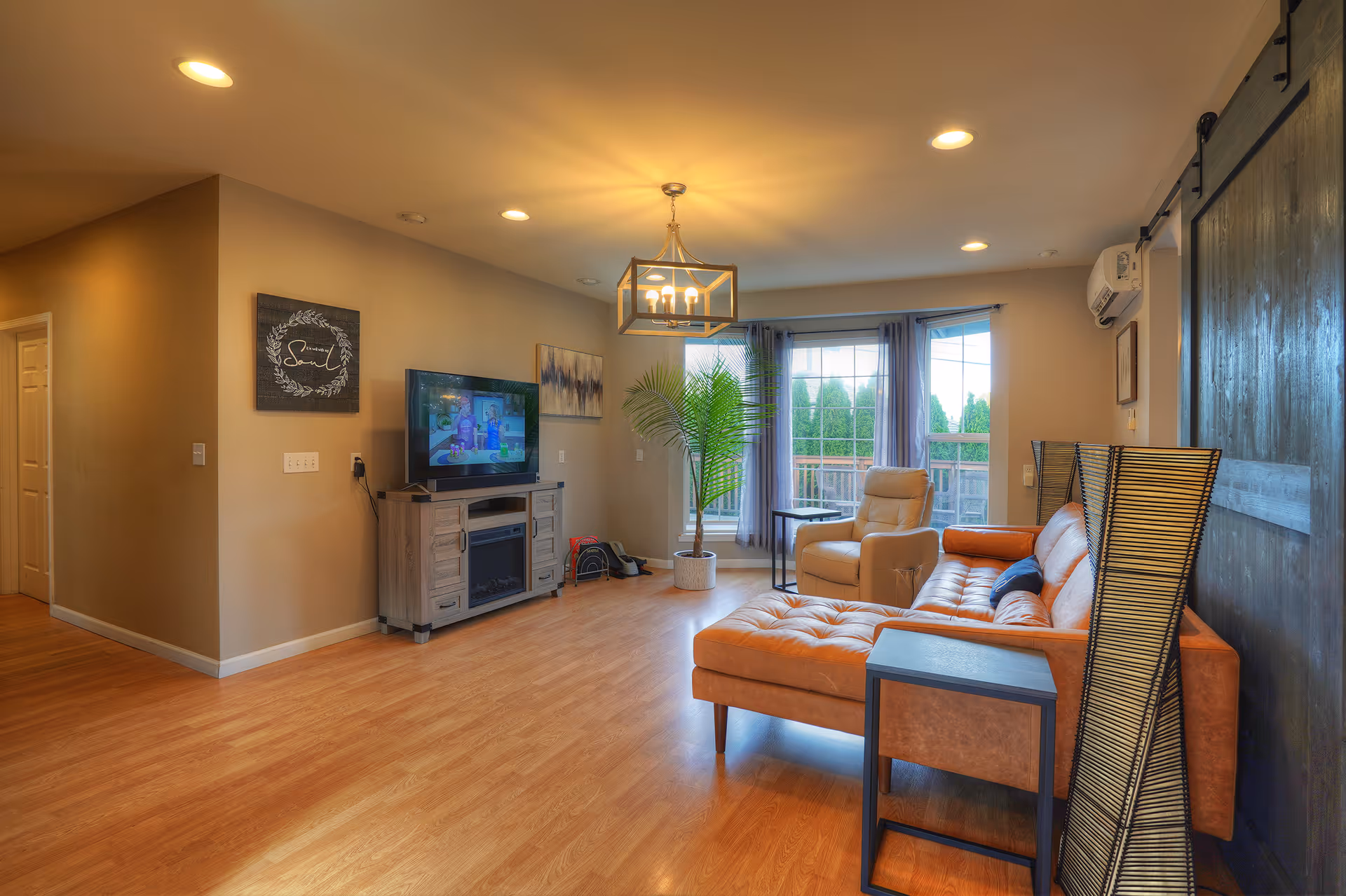 A cozy living room with wooden flooring, a tan leather sectional sofa, a beige recliner chair, a TV on a wooden stand, a potted plant near large windows with sheer curtains, and a modern ceiling light fixture.