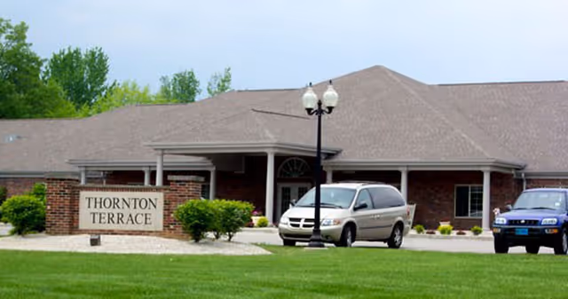 Exterior view of Thornton Terrace Health Campus building with a brick facade, a covered entrance, two parked vehicles, a street lamp, and a sign reading 'Thornton Terrace' on a landscaped lawn.