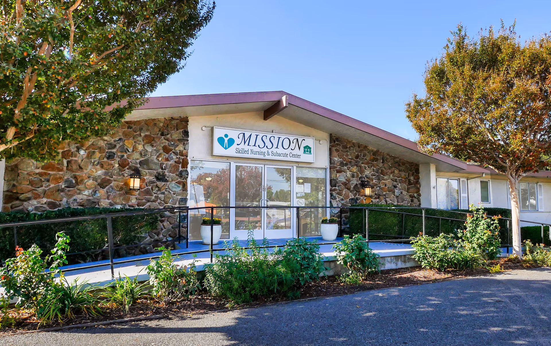 Exterior front view of Mission Skilled Nursing & Subacute Center building with stone facade, glass double doors, a ramp with railings, potted plants, and trees on a sunny day.