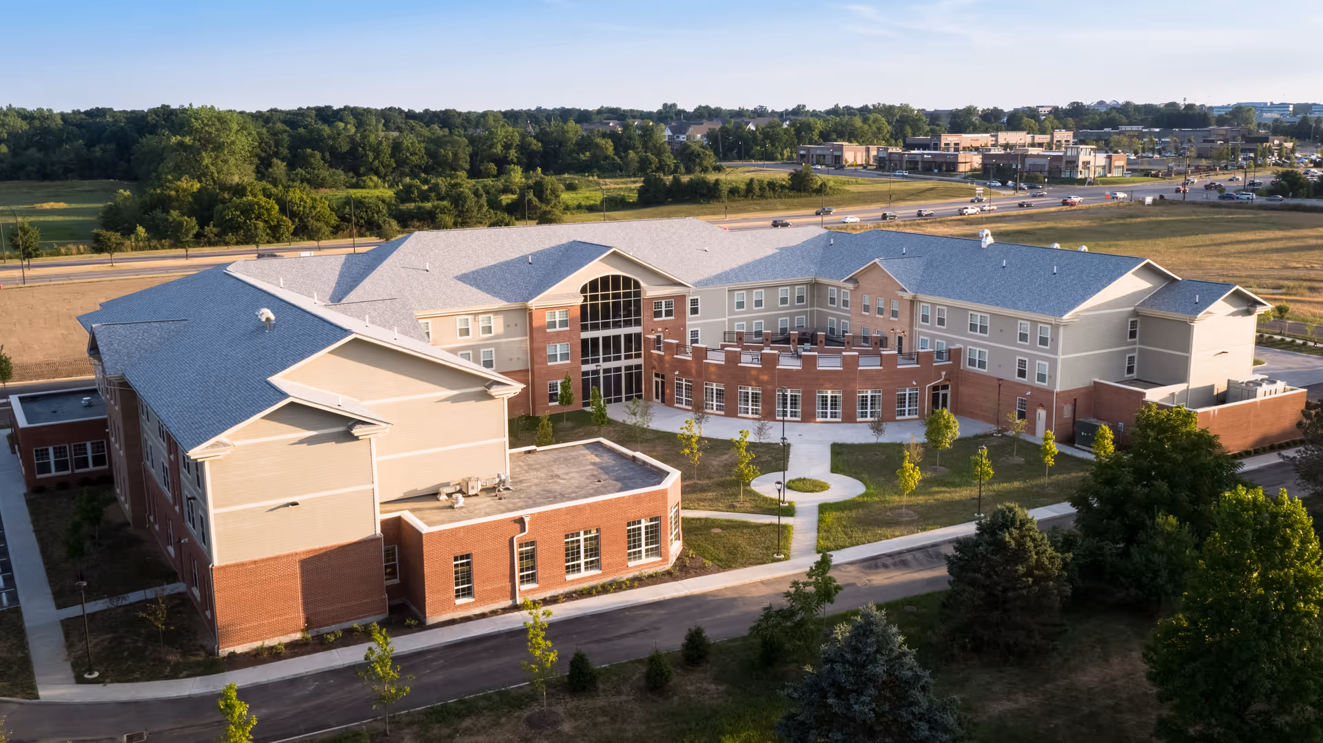 Aerial view of a large, modern senior living facility building with a combination of brick and beige siding exterior. The building is surrounded by landscaped green areas, young trees, and paved walkways. In the background, there is a road with cars and a commercial area with shops and buildings.