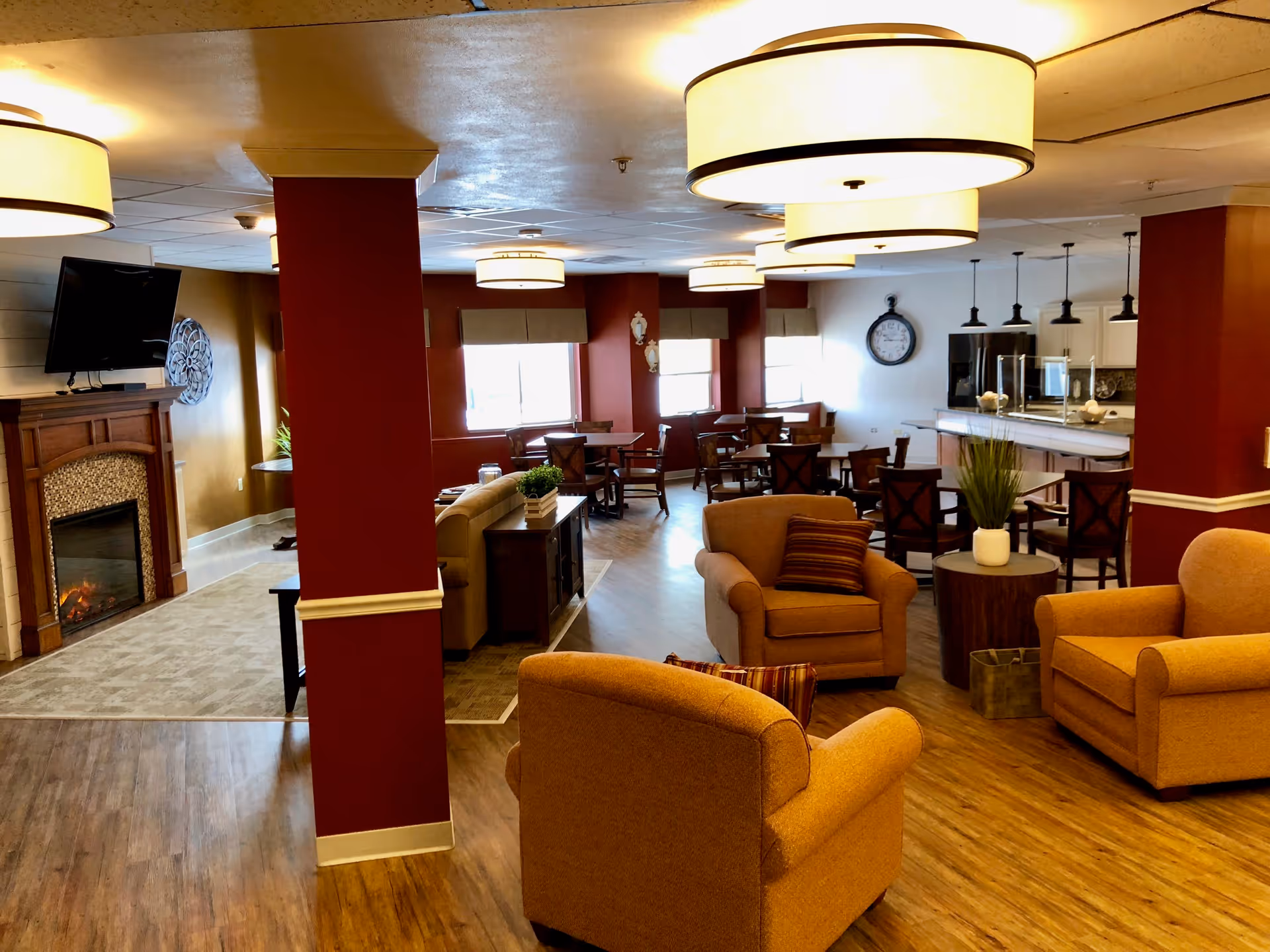 A cozy senior living common area featuring comfortable armchairs with striped pillows, a fireplace with a mounted TV above it, several round tables with chairs, and a kitchen area with bar stools and pendant lighting. The walls are painted in warm tones with large windows letting in natural light.
