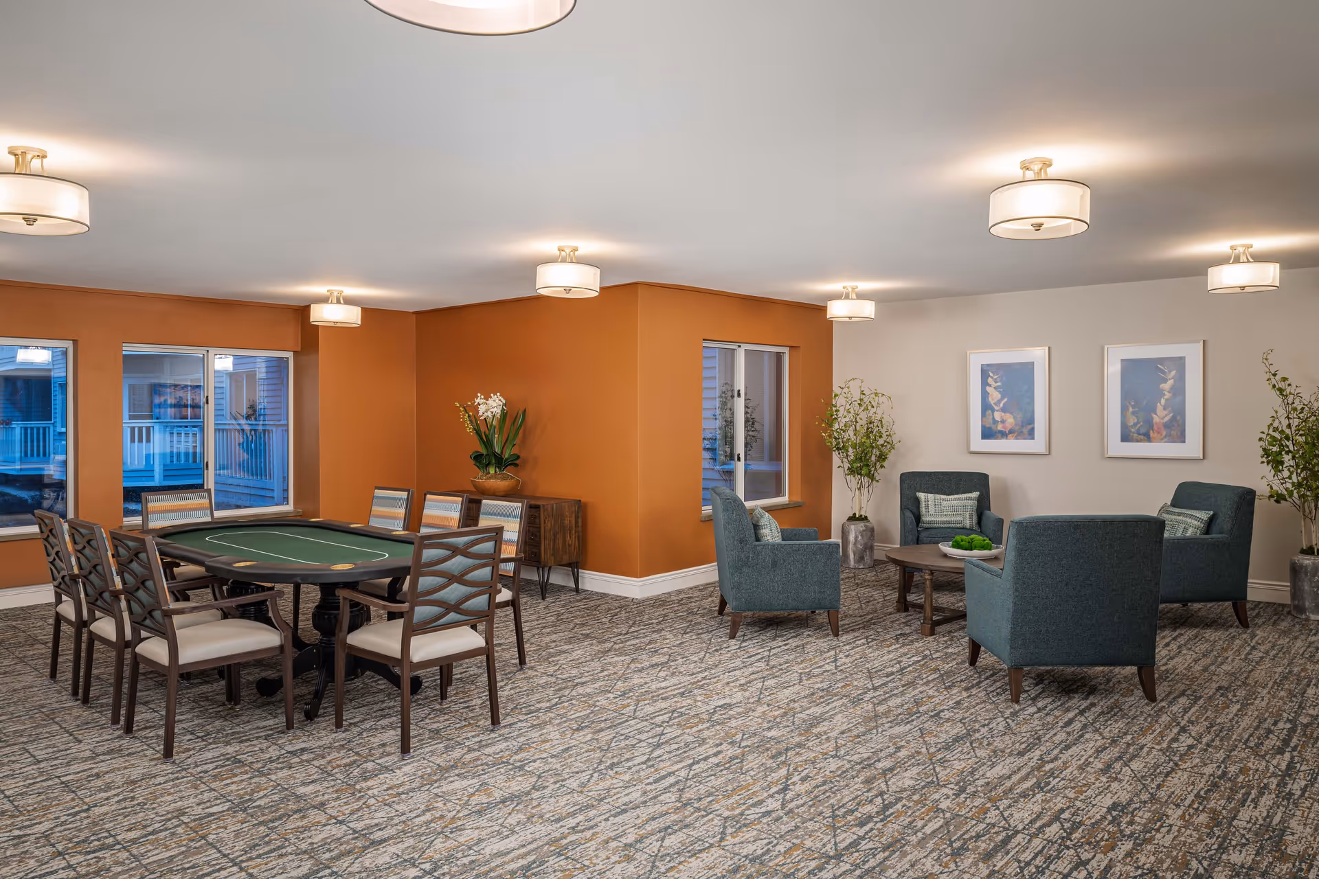 A spacious senior living common area with a card table surrounded by eight chairs on the left and a seating area with four blue armchairs around a round wooden coffee table on the right. The walls are painted orange and white, with two framed artworks and potted plants. The room is well-lit with ceiling lights and has large windows.