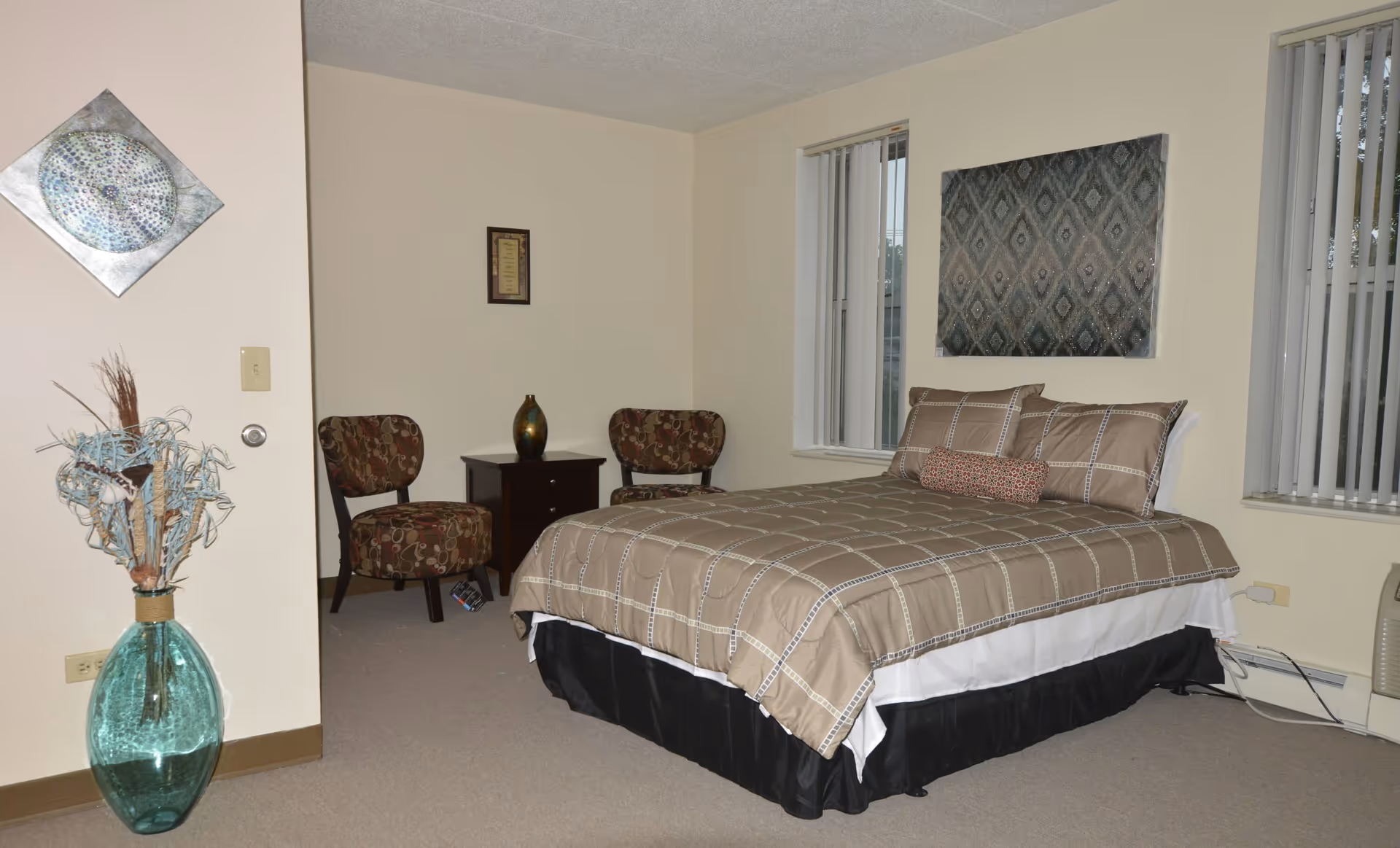 A bedroom with a neatly made bed featuring beige and brown checkered bedding, two patterned chairs with a small dark wooden table between them, a decorative vase on the table, two windows with vertical blinds, a large decorative wall hanging above the bed, and a large green glass vase with dried plants in the corner.