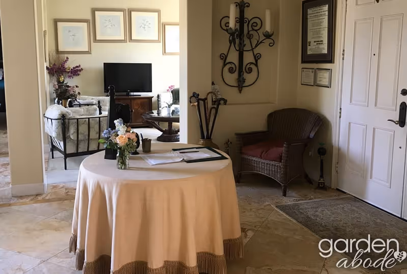 Interior view of a senior living facility entrance area with a round table covered with a beige tablecloth and a flower vase on top. To the right is a wicker chair with a red cushion and a wall-mounted decorative candle holder. In the background, there is a living room area with a TV, framed pictures on the wall, and cushioned chairs.