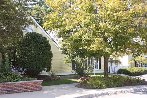 Exterior view of a senior living facility with light yellow siding, surrounded by green trees and bushes. A brick planter is visible on the left side near the sidewalk.