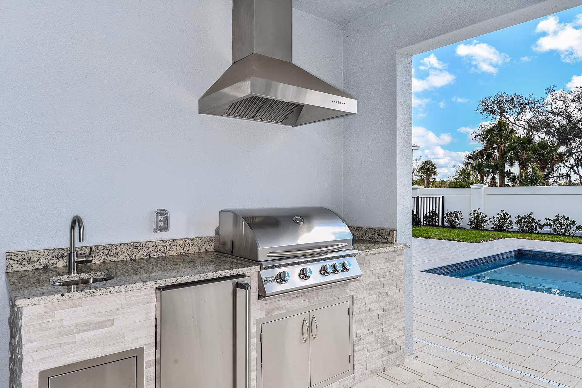 Outdoor kitchen area with a stainless steel grill, range hood, small sink, and granite countertop. The area opens to a patio with a swimming pool, surrounded by a white fence and greenery under a partly cloudy sky.