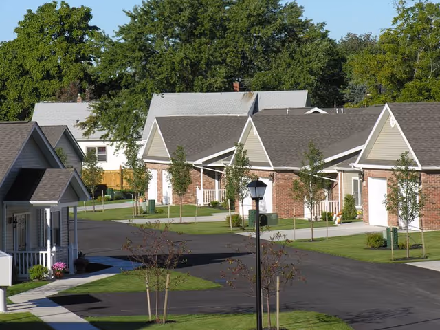 View of a residential community with single-story houses featuring brick and siding exteriors, driveways, small trees, and well-maintained lawns under a clear sky.