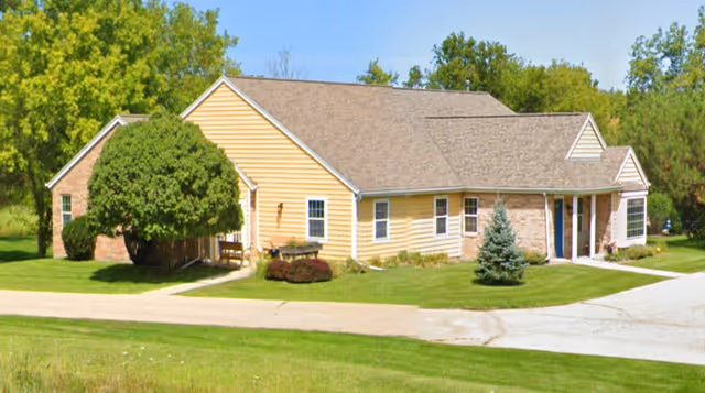 Single-story building with yellow siding and a brown shingled roof surrounded by green grass, trees, and shrubs under a clear blue sky.