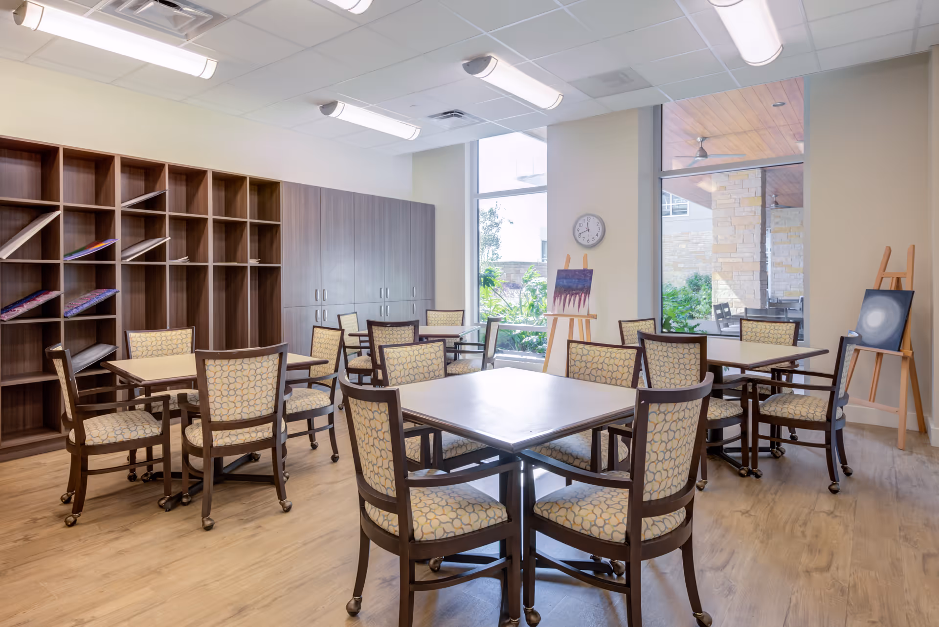 A bright activity room with several square tables surrounded by cushioned chairs on wheels. The room has wooden flooring, large windows letting in natural light, and a wall clock. There are wooden shelves and cabinets along one wall, with some paintings displayed on easels near the windows. Outside the windows, greenery and part of a stone building are visible.