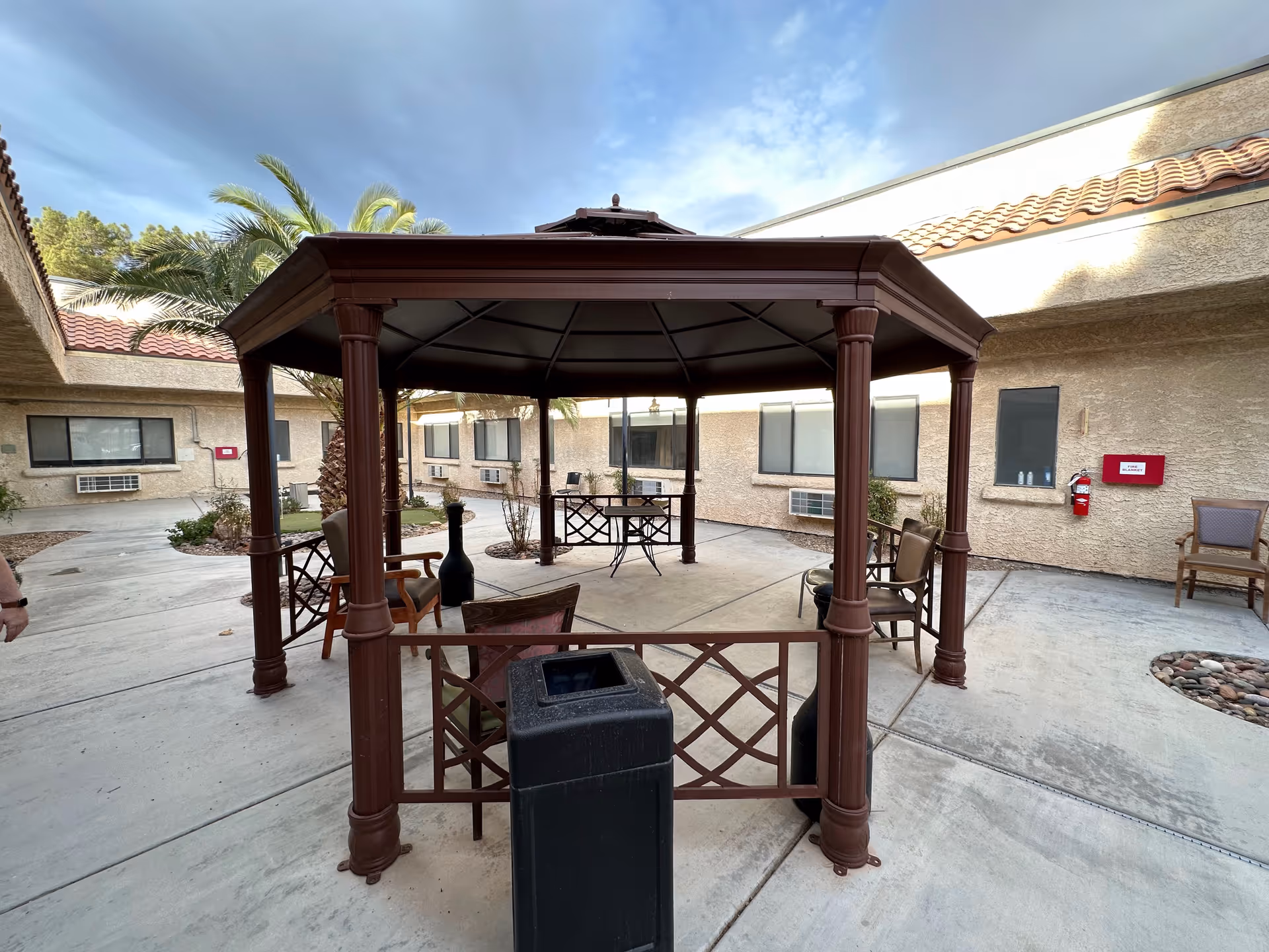 Outdoor courtyard area at Mission Pines Nursing & Rehabilitation Center featuring a brown gazebo with several chairs arranged underneath it. Surrounding the gazebo are beige stucco building walls with windows and air conditioning units. There are palm trees and a clear sky with some clouds visible above.