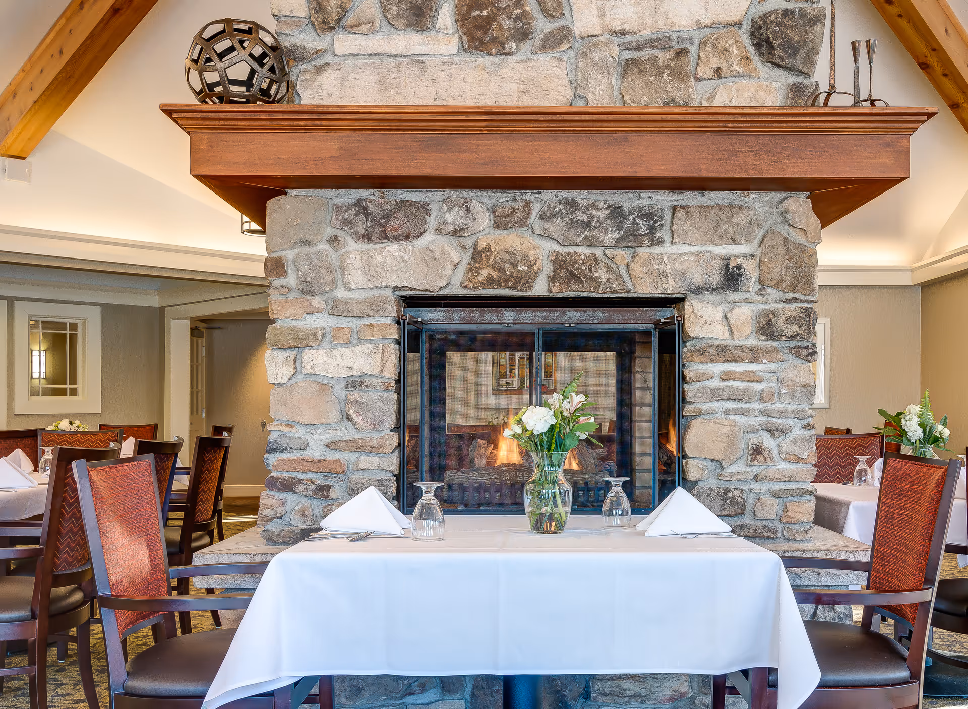 A dining area with a stone fireplace in the center. The fireplace has a wooden mantel with decorative items on top. In front of the fireplace is a table covered with a white tablecloth, set with folded white napkins, upside-down glasses, and a vase with white and pink flowers. Surrounding the table are wooden chairs with red upholstery. Additional tables and chairs are visible in the background.