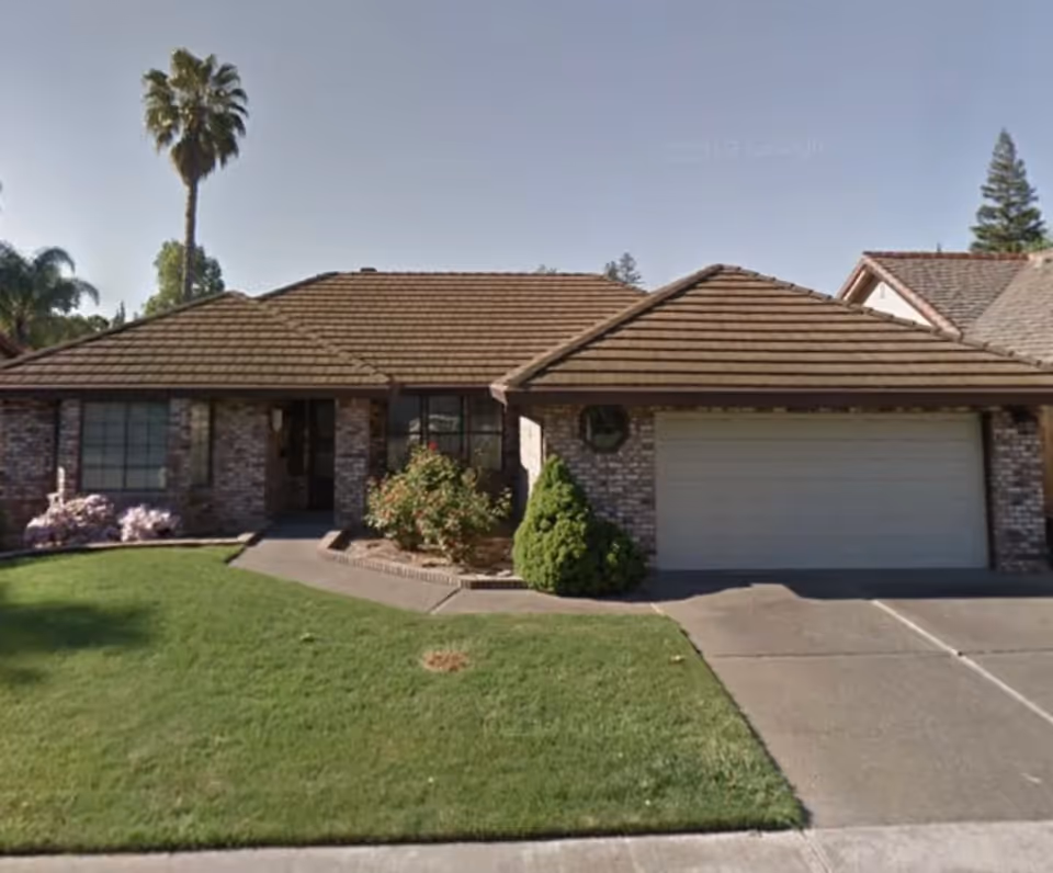 Single-story brick house with a brown tiled roof, a two-car garage, a well-maintained green lawn, and various shrubs and a tall palm tree in the front yard under a clear sky.