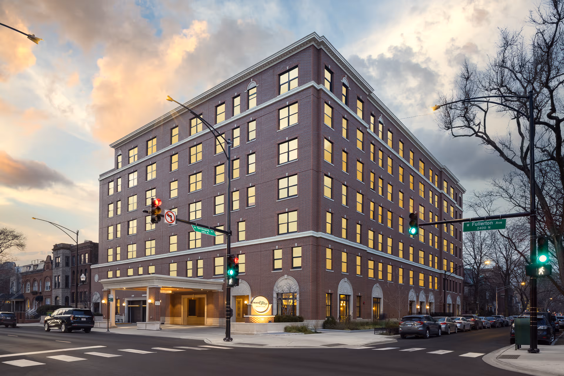Exterior view of a multi-story brick building at dusk with many windows lit from inside. The building is located at the corner of two streets with traffic lights and street signs visible. Several parked cars line the street, and leafless trees are seen on the sidewalk.