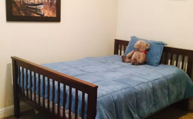 A bedroom with a wooden bed frame, a blue comforter, a matching blue pillow, and a teddy bear placed on the bed. A framed picture hangs on the wall above the bed.