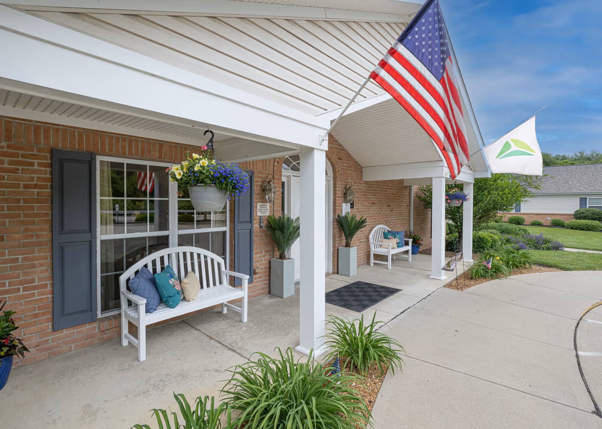 Covered entrance area of a brick building with two white benches adorned with colorful pillows. Hanging flower baskets and potted plants decorate the space. Two flags, one American and one with a green and blue logo, are mounted on the building. The walkway curves around the entrance, and there is a well-maintained lawn and shrubbery in the background.