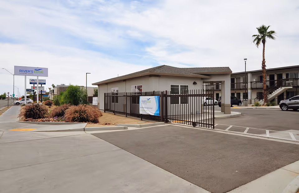 Exterior view of River's Edge assisted living facility showing a gated entrance, a single-story building, a two-story building with balconies, a palm tree, and a sign with the facility's name along a street with some cars parked nearby.