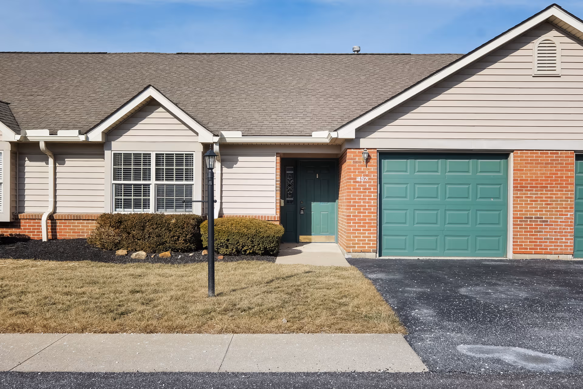 Exterior view of a single-story residential unit with beige siding and red brick accents. The unit features a green front door, a green garage door, a window with white blinds, a small front lawn with bushes, a sidewalk, and a black lamp post in front.