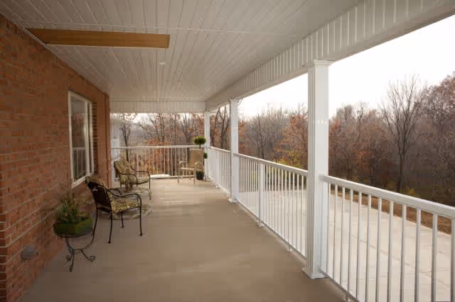 A spacious covered porch with a white ceiling and railing, two metal chairs with cushions, and potted plants. The porch overlooks a wooded area with leafless trees.