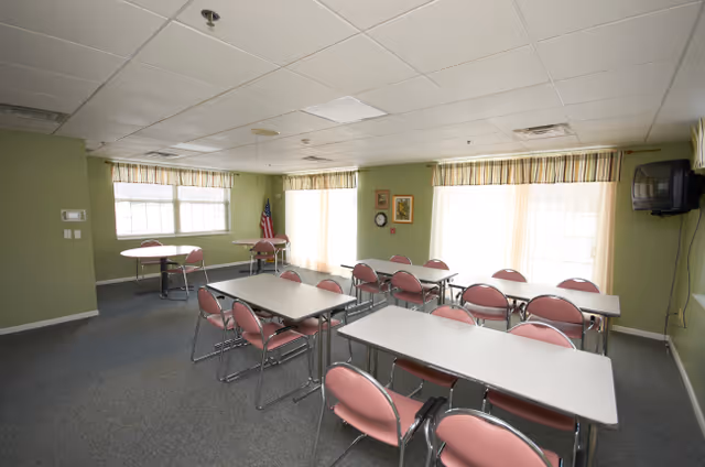 A well-lit room with multiple rectangular tables and pink cushioned chairs arranged around them, large windows with striped curtains allowing natural light, an American flag in the corner, framed pictures on the wall, and a wall-mounted television.
