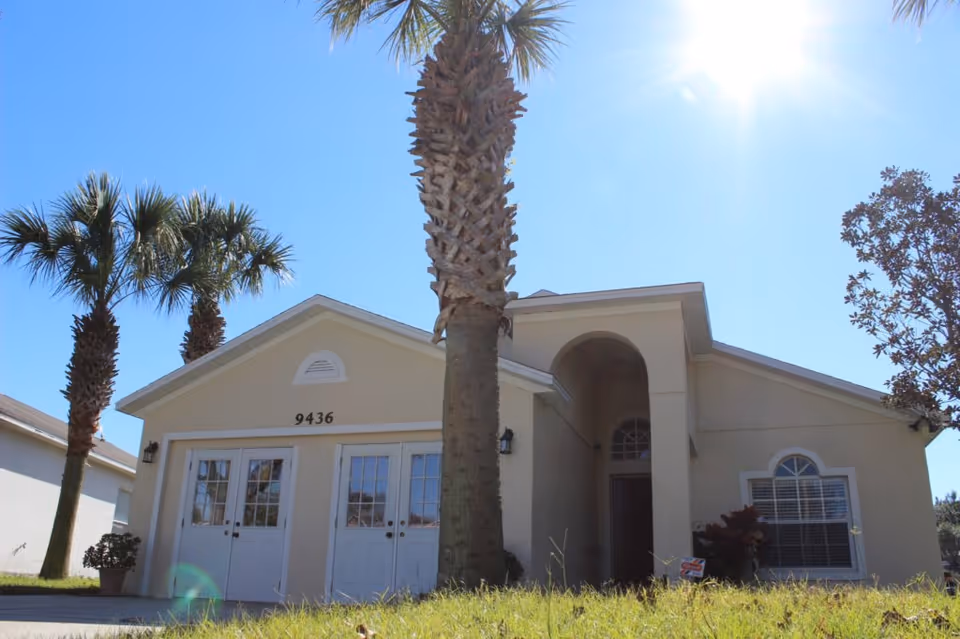 Exterior view of a single-story beige building with the number 9436 above two white double doors. There are palm trees and a small tree in front of the building, with bright sunlight and a clear blue sky.