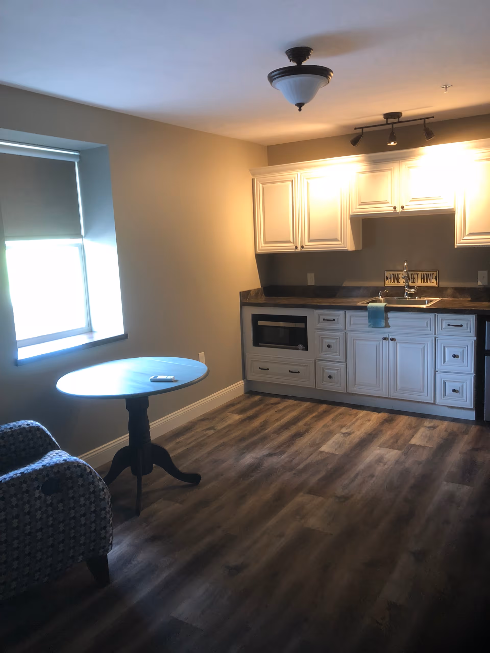 A small kitchen area with white cabinets, a countertop, a sink, and a microwave built into the lower cabinets. There is a round wooden table with a remote control on it and a patterned armchair next to the window with a closed blind. The floor is wood-style laminate, and the walls are painted beige. A ceiling light fixture and track lighting illuminate the space.