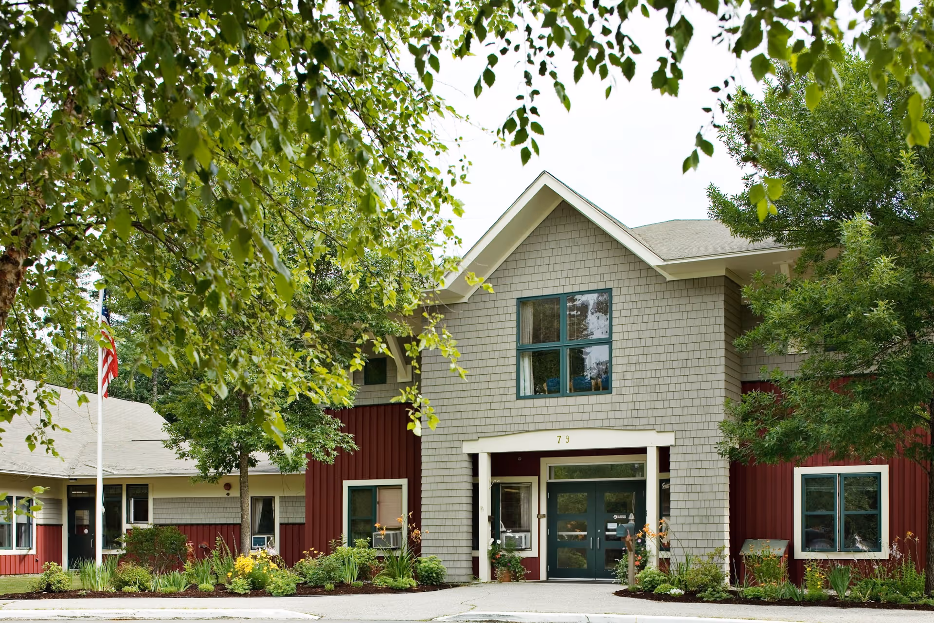 Exterior view of a senior living facility building with gray shingles and red siding, surrounded by green trees and plants. The entrance has double doors with the number 79 above them, and an American flag is visible on the left side.