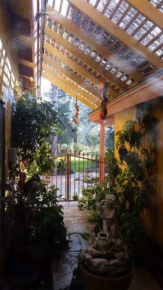 A covered outdoor patio area with a wooden pergola roof, lush green plants, and a small stone statue of a cherub holding a pitcher. The area opens to a garden with a metal gate and trees in the background. Sunlight filters through the pergola, creating a serene and inviting atmosphere.