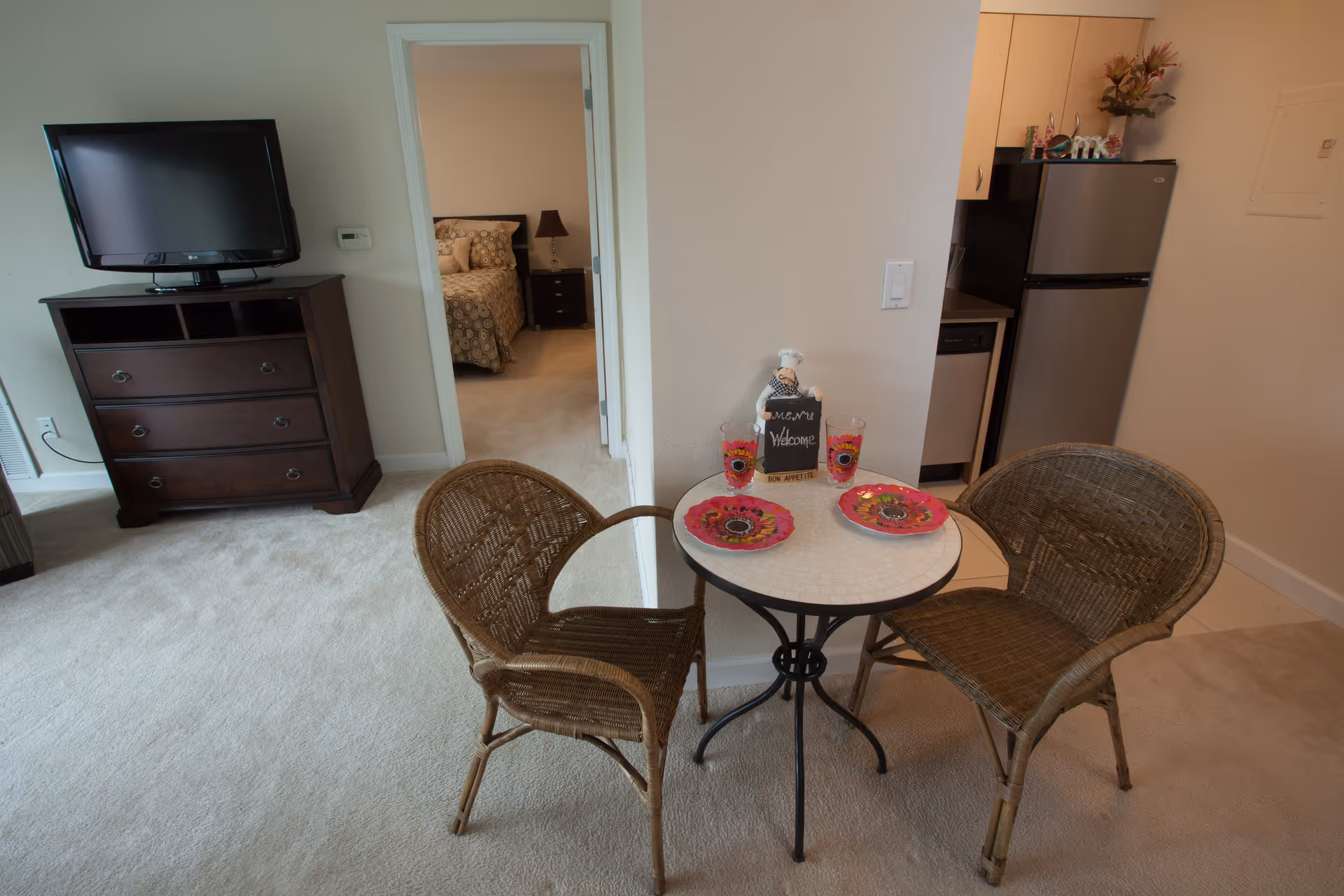 Interior view of a senior living facility apartment showing a small round dining table with two wicker chairs, set with two colorful plates and glasses, and a decorative chef figurine holding a welcome menu sign. In the background, there is a dark wooden dresser with a flat-screen TV on top, a doorway leading to a bedroom with a bed and nightstand, and a partial view of a kitchen area with a refrigerator and dishwasher.