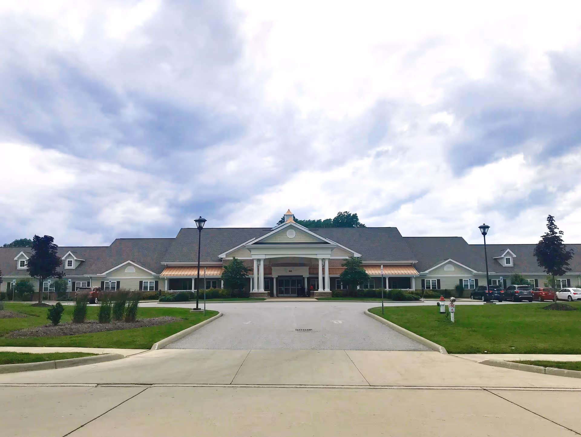 Front exterior view of Highland Pointe Health & Rehabilitation Center, a large single-story building with a peaked roof, white columns at the entrance, and a driveway leading up to the main doors. The sky is cloudy and there are some trees and parked cars visible on the sides.