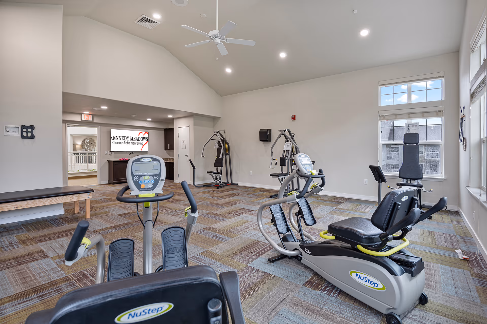 A bright fitness room in a senior living facility with exercise equipment including NuStep recumbent cross trainers and weight machines. The room has large windows letting in natural light, a ceiling fan, and a patterned carpet. A sign for Kennedy Meadows Gracious Retirement Living is visible in the background.
