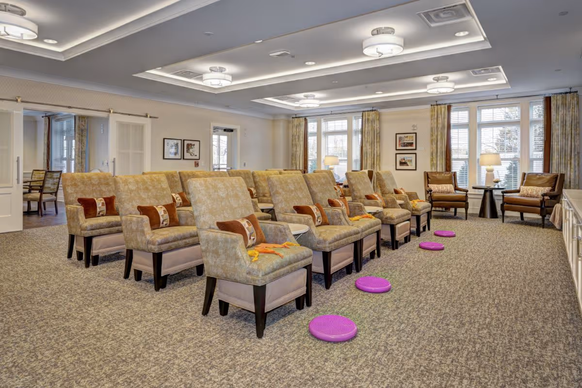 A spacious, well-lit common area with multiple rows of beige upholstered chairs, each with a small pillow and a yellow towel. Purple balance cushions are placed on the carpeted floor in front of the chairs. The room has large windows with curtains, two armchairs with pillows near the windows, and framed pictures on the walls. The ceiling features recessed lighting and decorative light fixtures.