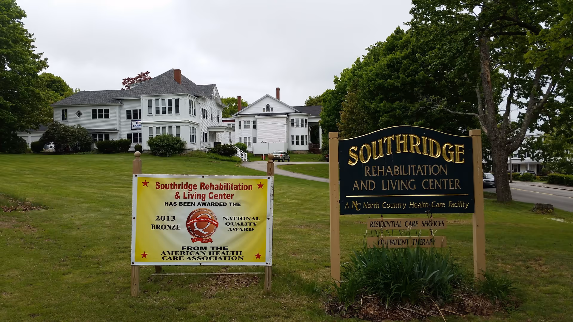 Exterior view of Southridge Rehabilitation and Living Center, showing a large white building with multiple windows and a green lawn. Two signs are visible in the foreground: one black and gold sign with the facility name and services offered, and another yellow sign announcing a 2013 Bronze National Quality Award from the American Health Care Association.