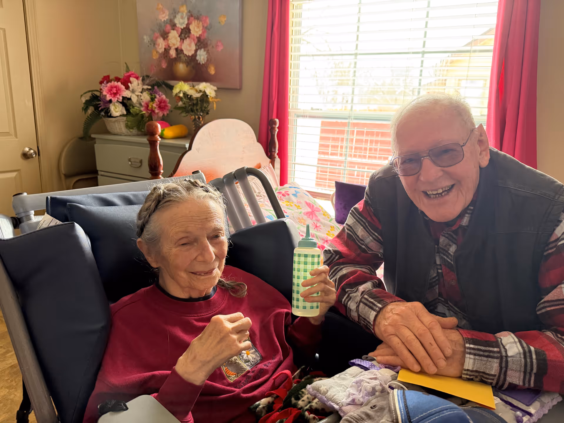 An elderly woman sitting in a wheelchair holding a green and white checkered bottle, smiling gently. Next to her is an elderly man wearing glasses and a plaid shirt with a black vest, smiling warmly. They are in a cozy room with a window covered by blinds and red curtains, a wooden bed with floral bedding, a dresser with flower arrangements, and a painting of flowers on the wall.