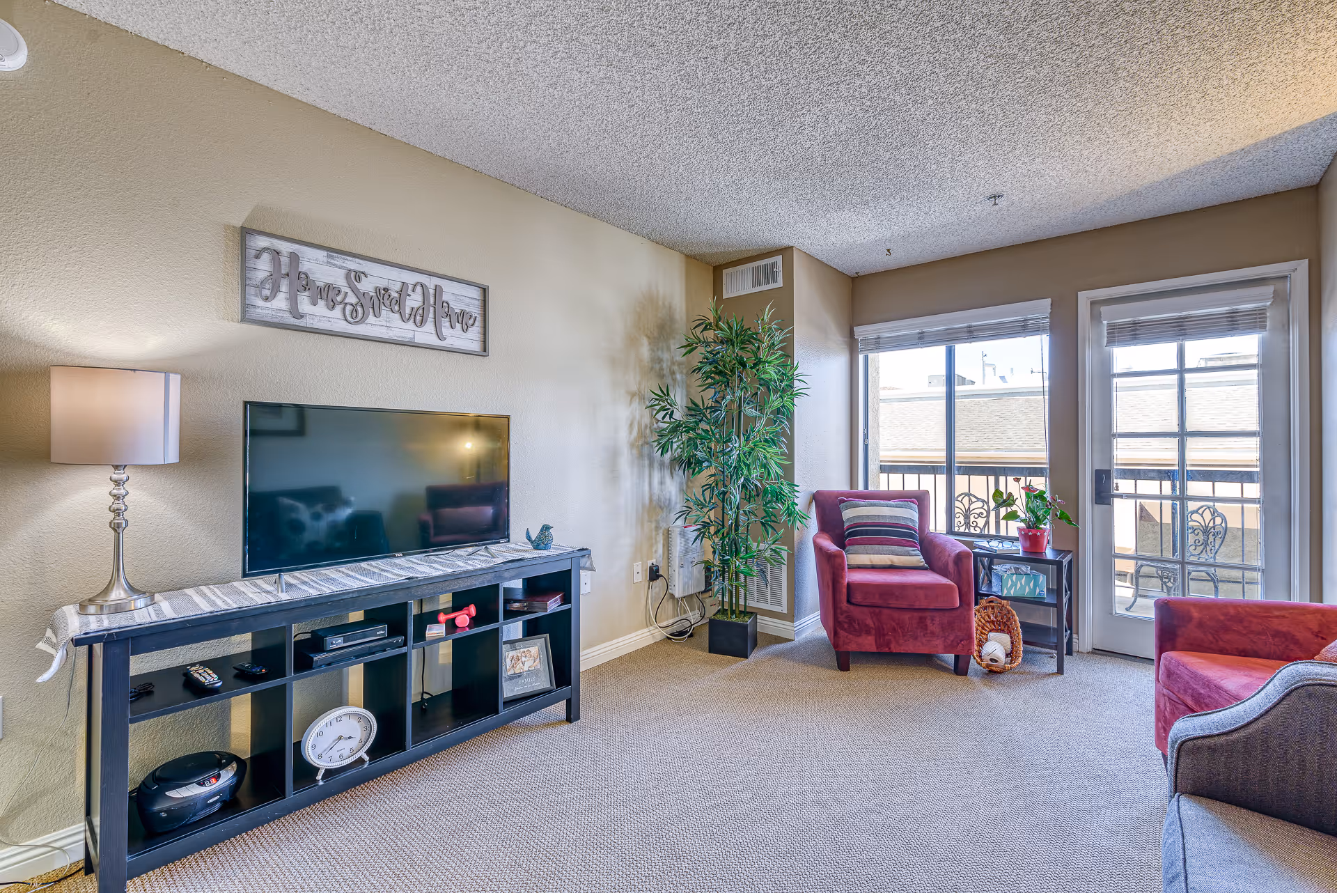 A cozy living room with beige walls and carpet. There is a black TV stand with a flat-screen TV, a table lamp, and various decorative items including a clock and a framed photo. Two red armchairs with striped cushions are positioned near a window and glass door leading to a balcony with outdoor seating. A tall green plant is placed in the corner next to the window. Above the TV, a wall sign reads 'Home Sweet Home.'