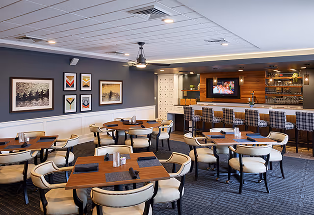 A cozy dining area with several wooden tables and cushioned chairs arranged neatly. The tables are set with placemats, napkins, and salt and pepper shakers. On one side, there is a bar counter with high plaid upholstered stools and a TV mounted on a wooden accent wall behind the bar. The walls are decorated with framed artwork and the ceiling has recessed lighting and a ceiling fan.