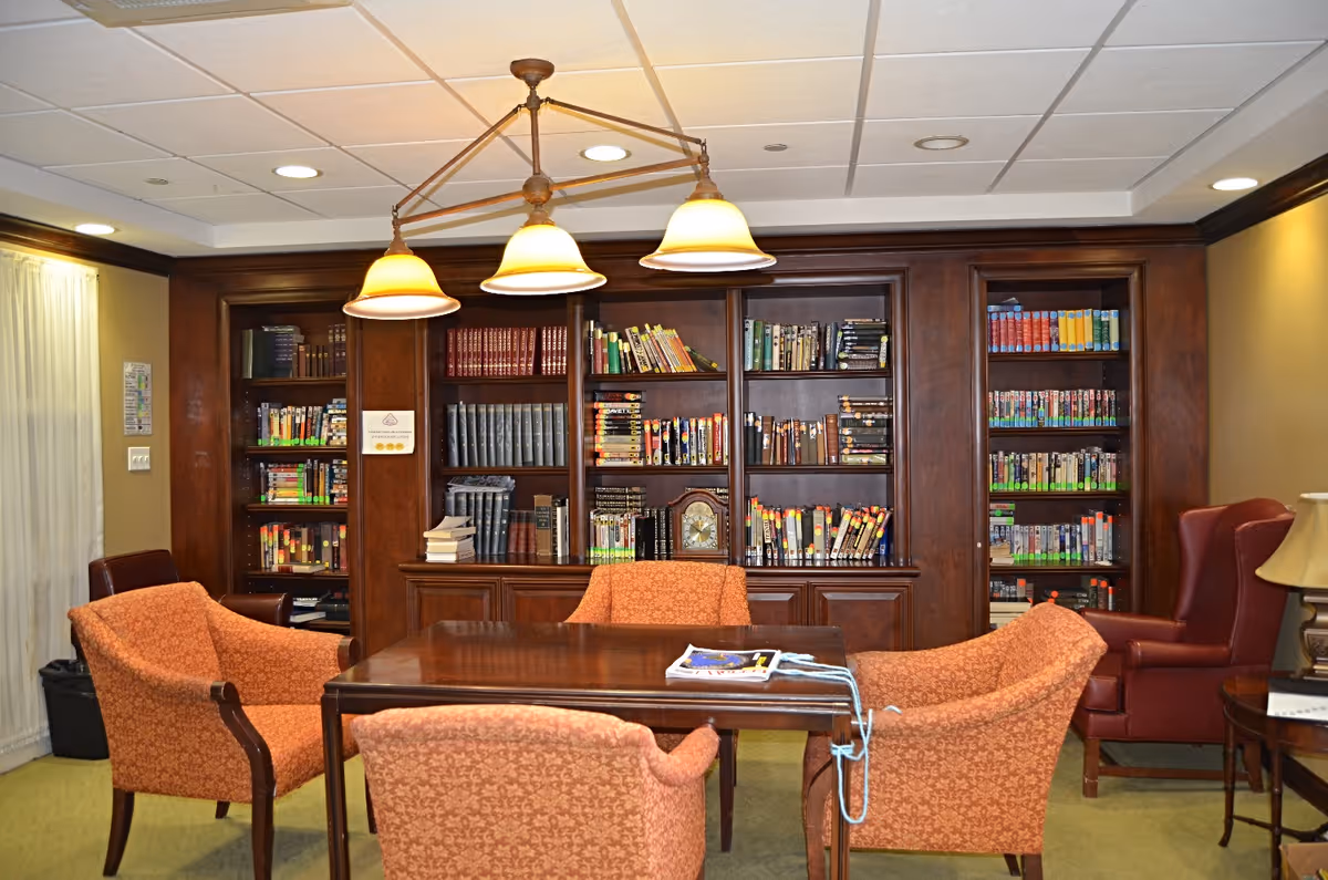 A cozy library or reading room with a wooden table surrounded by four orange upholstered chairs. Behind the table is a large wooden bookshelf filled with books and some decorative items. A three-light ceiling fixture hangs above the table. To the right, there is a red leather armchair next to a small side table with a lamp. The room has a warm and inviting atmosphere.
