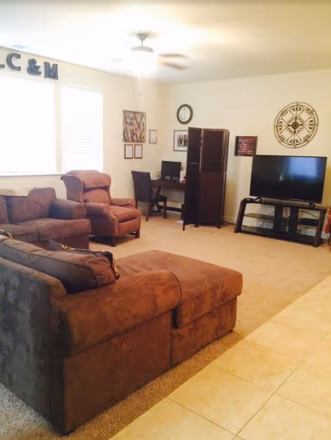 A cozy living room with a brown sectional sofa, two armchairs, a TV on a stand, a small desk with a computer, wall decorations including a clock and framed pictures, and a ceiling fan. The room has carpeted flooring and a tiled area in the foreground.