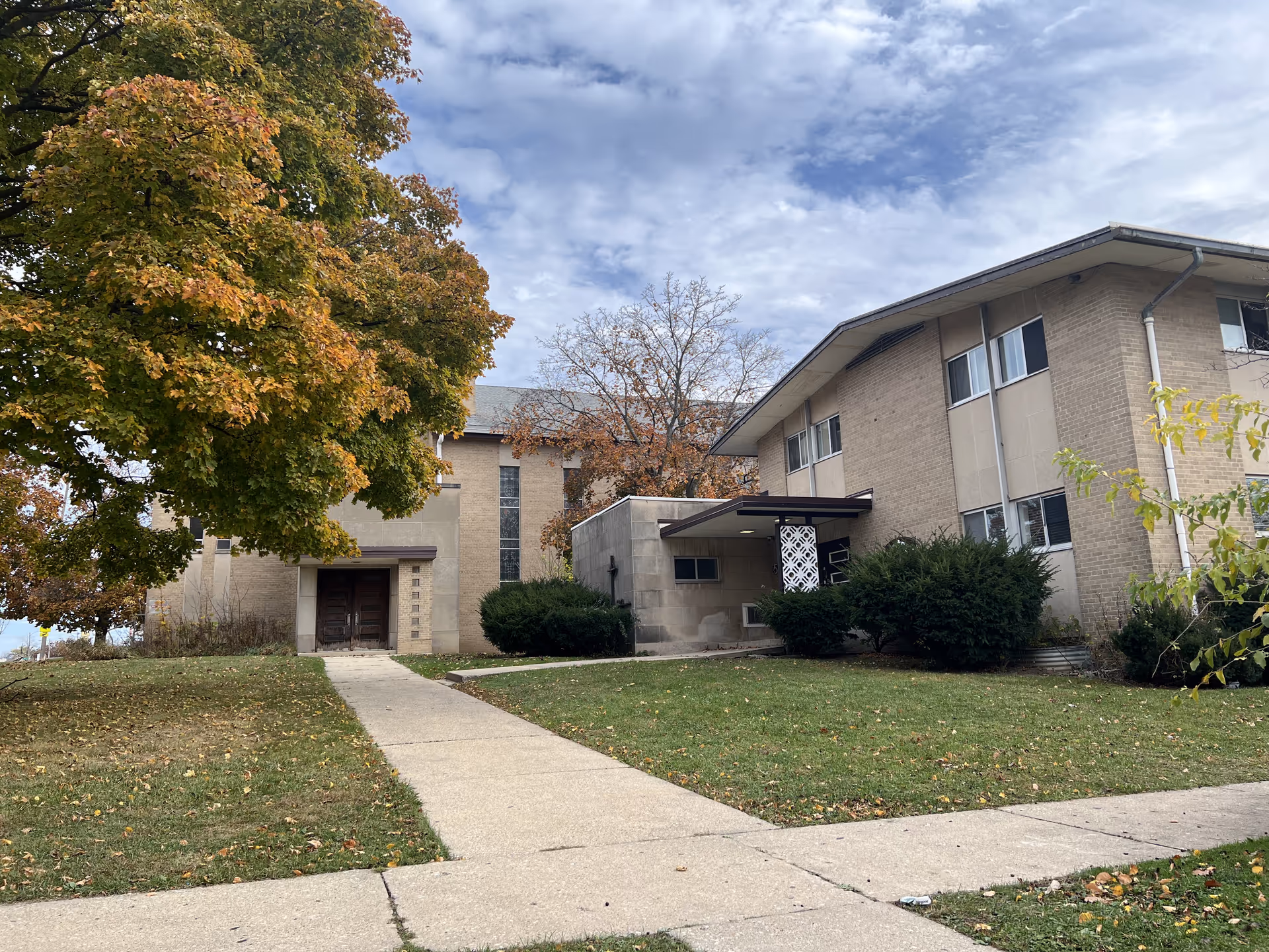Exterior view of a two-story brick building with a sidewalk leading to the entrance. There are large trees with autumn-colored leaves and green bushes near the building. The sky is partly cloudy.