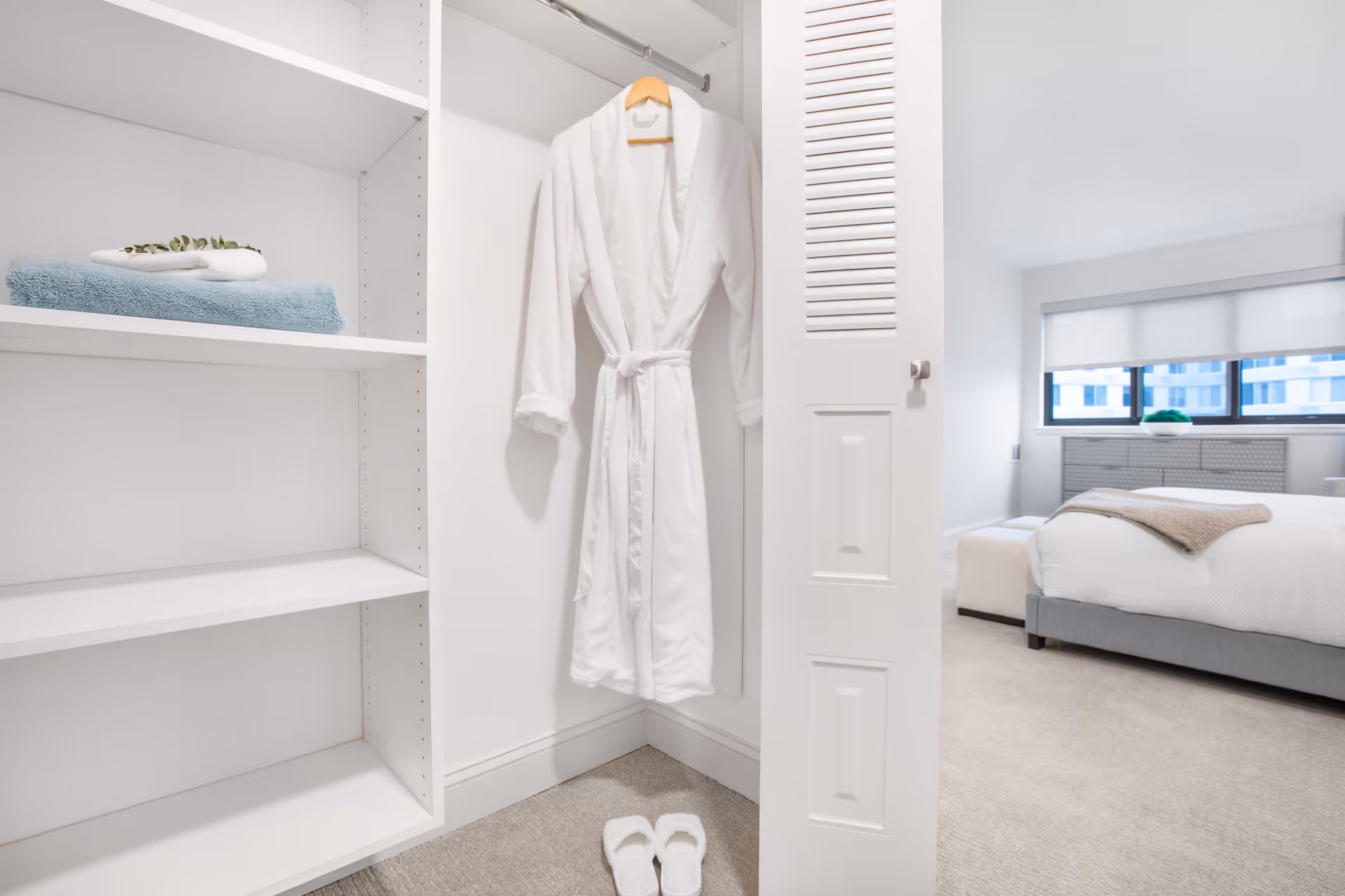 A white bathrobe hanging inside an open closet with white shelves, a pair of white slippers on the floor, and a view into a bright bedroom with a bed, ottoman, and large windows with blinds.