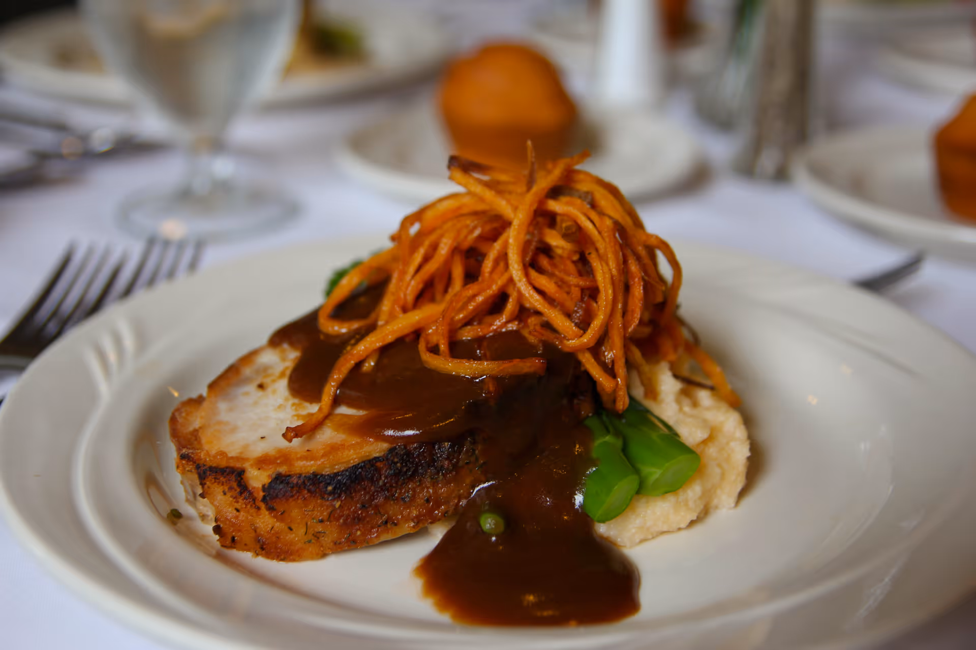A plated meal featuring a slice of roasted meat topped with brown gravy and crispy fried thin potato strings, served with green beans and mashed potatoes on a white plate with a white tablecloth and blurred background of other plates and glassware.