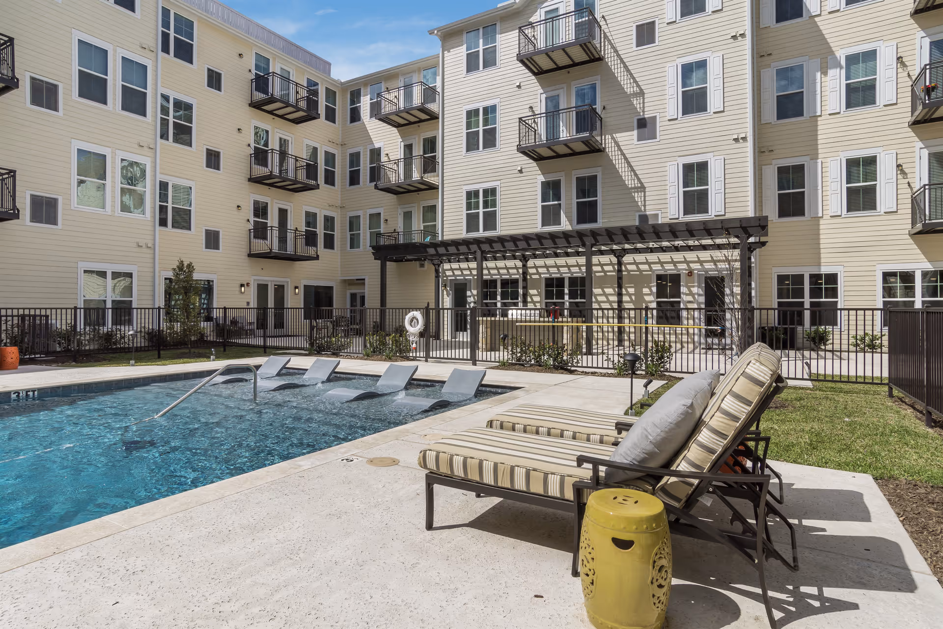 Outdoor swimming pool area at Park Creek Active Retirement facility with lounge chairs and a yellow side table on the pool deck, surrounded by a multi-story residential building with balconies and windows.