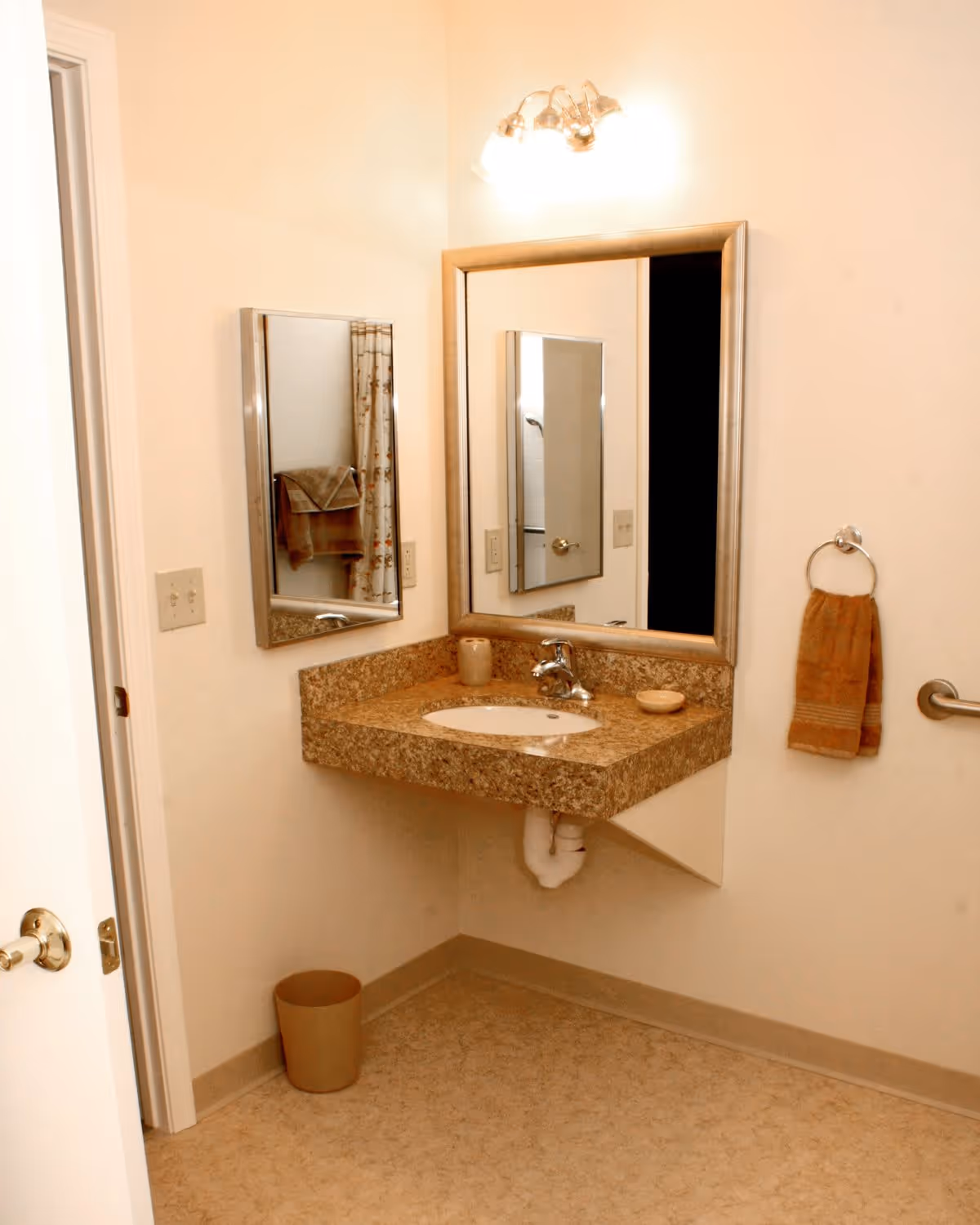 A bathroom corner with a granite countertop sink mounted on the wall, a large rectangular mirror above the sink, a smaller mirror on the adjacent wall, a towel ring with a brown towel, a beige trash bin on the floor, and a light fixture above the large mirror.