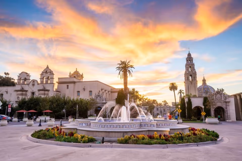 A scenic outdoor plaza at sunset featuring a large circular fountain with water jets surrounded by colorful flowers and greenery. Historic Spanish-style buildings with ornate architectural details and a tall bell tower are visible in the background under a vibrant sky with orange and purple clouds.