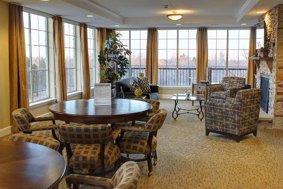 A bright and spacious living room area with large windows letting in natural light. The room features a round wooden table surrounded by patterned upholstered chairs, a seating area with a sofa and two armchairs, a glass coffee table, and a stone fireplace on the right. There are beige curtains on the windows and a potted plant near the sofa.