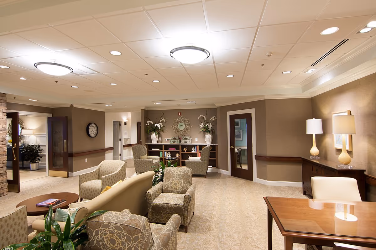 A well-lit, spacious common area with comfortable armchairs and sofas arranged around a coffee table. The room features beige walls, carpeted floors, decorative plants, a wall clock, and a wooden cabinet with two lamps. There are doors leading to other rooms and a bookshelf with decorative items and flowers.